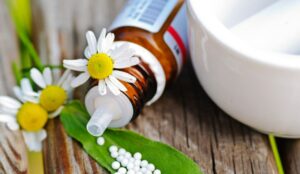 Homöopathie-Fläschchen liegend, neben einem Mörser, Kamilleblüten und Globuli auf eiunem Blatt.